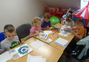 Pięcioro dzieci siedzi przy stole i dokańcza malowanie płótna.